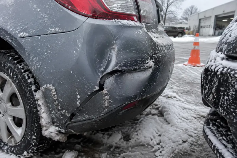 A cracked and dented rear bumper cover on a compact car in a snowy parking lot, with road salt on the paint and an orange cone nearby