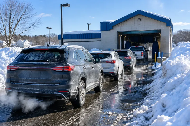 Line of salt-dusted cars waiting for an express car wash tunnel the morning after a snowstorm, with high plowed snowbanks along the lane