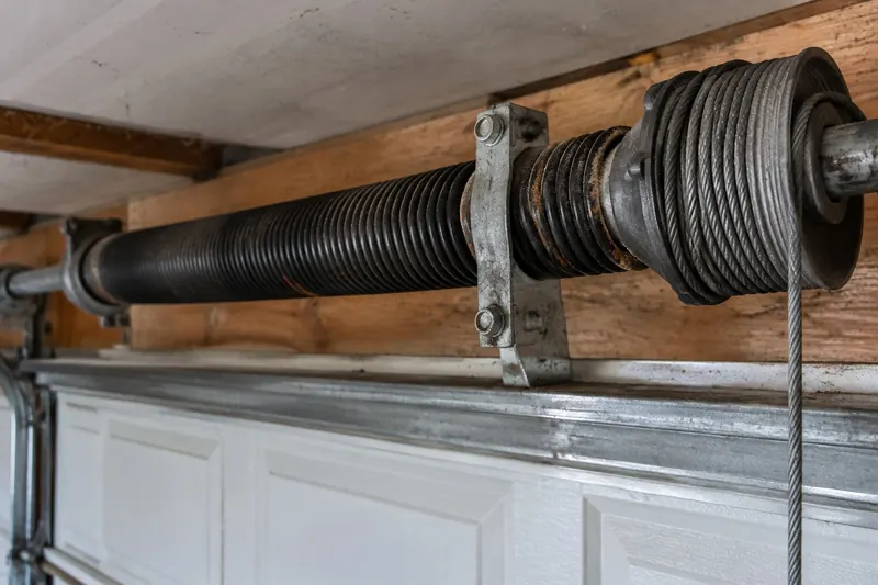 Close view of a black steel torsion spring on its shaft with a cable drum and center bracket above a garage door