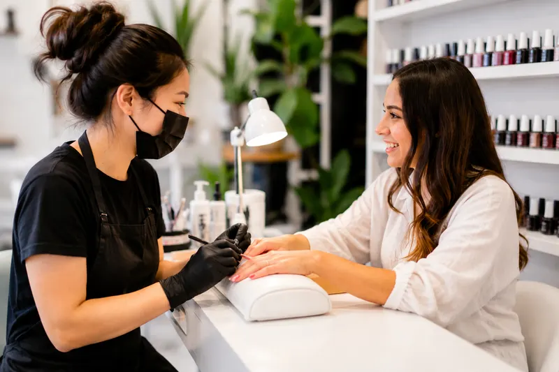 Nail technician in a mask filing a client's nails at a white station with a polish wall and plants behind
