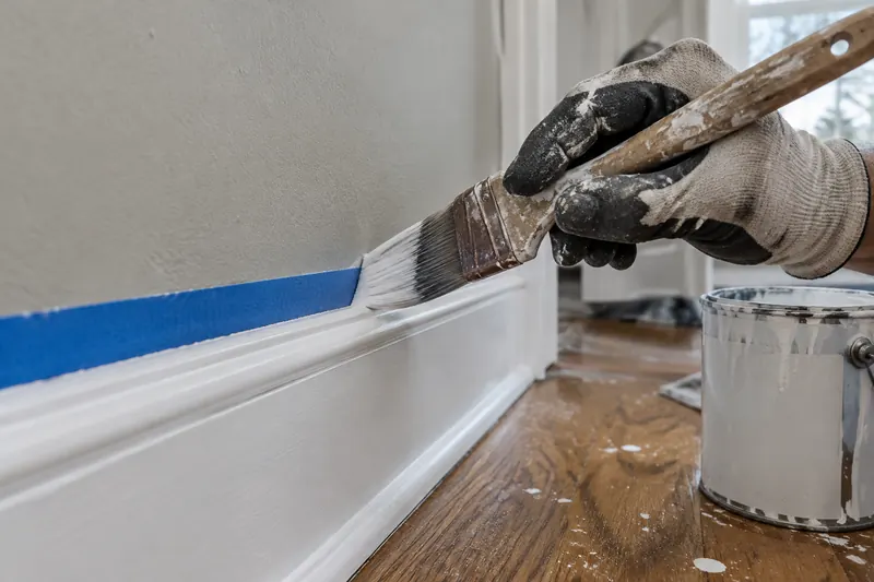 A gloved hand cutting in a crisp line with an angled brush along blue painters tape at the top of a freshly painted white baseboard