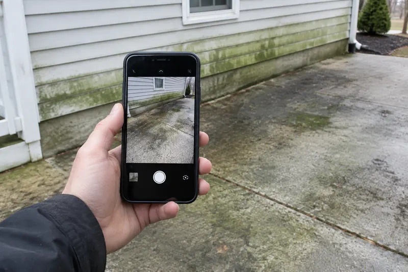 Homeowner hand holding a smartphone to photograph an algae-streaked concrete driveway and green-stained siding for a quote